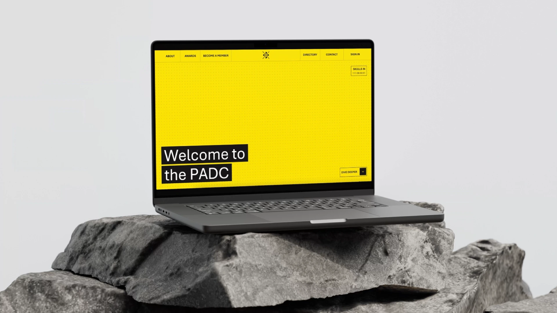 Yellow laptop on a rough stone surface displaying the PADC website with bold black and yellow branding